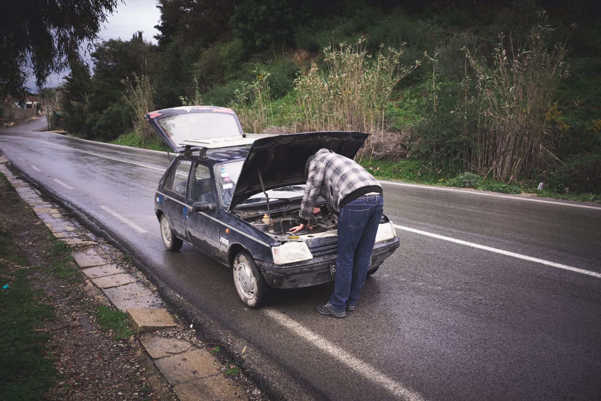 An individual repairing a car on a rainy road in Tabarka, showcasing urgent roadside vehicle maintenance.